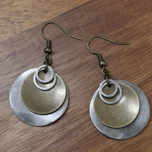 May include: A pair of dangle earrings featuring layered circular metal discs in brushed silver and antique brass tones. Each earring consists of a large silver disc, a smaller brass disc, and two tiny silver rings suspended from a dark metal hook.