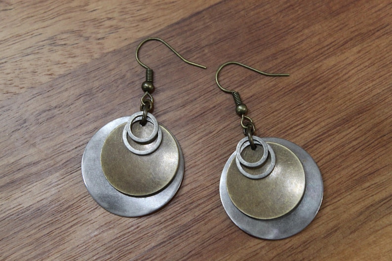 May include: A pair of dangle earrings featuring layered circular metal discs in brushed silver and antique brass tones. Each earring consists of a large silver disc, a smaller brass disc, and two tiny silver rings suspended from a dark metal hook.