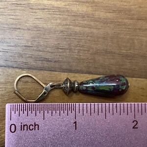 May include: A drop earring with a leverback closure and a metallic bead spacer attached to a teardrop-shaped bead featuring a mottled pattern of dark red, green, and black. A ruler placed underneath shows the earring measures approximately 1.5 inches.