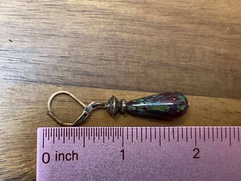 May include: A drop earring with a leverback closure and a metallic bead spacer attached to a teardrop-shaped bead featuring a mottled pattern of dark red, green, and black. A ruler placed underneath shows the earring measures approximately 1.5 inches.