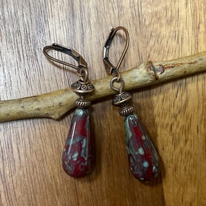 May include: A pair of drop earrings featuring teardrop-shaped beads with a mottled red and green glaze. The beads are suspended from antique copper-toned metal findings, including decorative spacer beads and secure lever-back ear wires.
