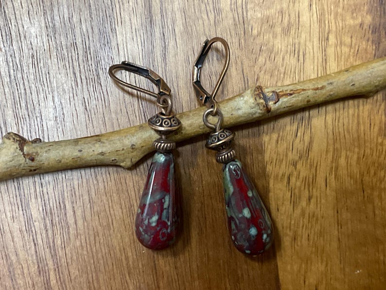 May include: A pair of drop earrings featuring teardrop-shaped beads with a mottled red and green glaze. The beads are suspended from antique copper-toned metal findings, including decorative spacer beads and secure lever-back ear wires.