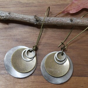 May include: A pair of dangle earrings featuring layered circular metal discs in brushed silver and antique brass tones. Each earring consists of a larger silver disc, a smaller brass disc, and two tiny silver rings suspended from a long brass hook.