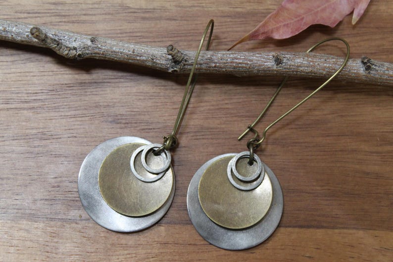 May include: A pair of dangle earrings featuring layered circular metal discs in brushed silver and antique brass tones. Each earring consists of a larger silver disc, a smaller brass disc, and two tiny silver rings suspended from a long brass hook.