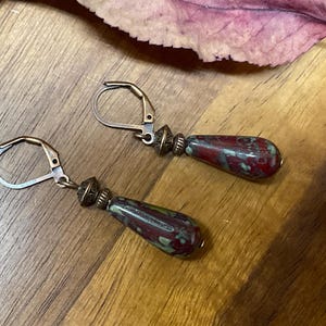 May include: A pair of drop earrings featuring teardrop-shaped beads with a mottled red and green pattern. The beads are attached to antique bronze-toned metal leverback hooks with small decorative metal spacers between the hook and the bead.