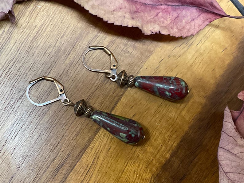 May include: A pair of drop earrings featuring teardrop-shaped beads with a mottled red and green pattern. The beads are attached to antique bronze-toned metal leverback hooks with small decorative metal spacers between the hook and the bead.
