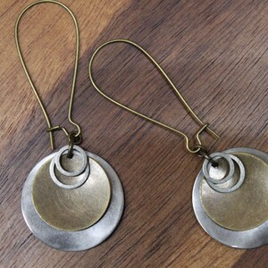 May include: A pair of dangle earrings featuring antique brass-toned ear wires. Each earring consists of a large silver-toned circular disc, a smaller brass-toned circular disc layered on top, and two small silver-toned rings attached to the center.