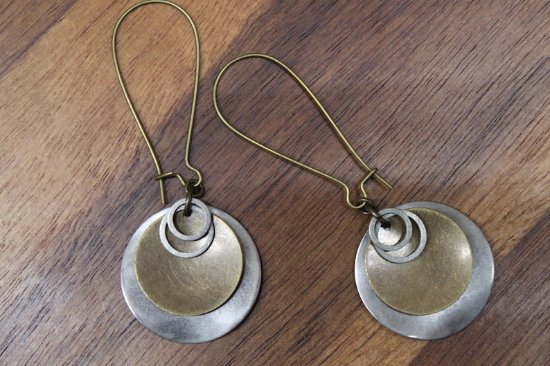 May include: A pair of dangle earrings featuring antique brass-toned ear wires. Each earring consists of a large silver-toned circular disc, a smaller brass-toned circular disc layered on top, and two small silver-toned rings attached to the center.