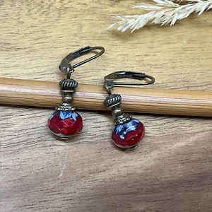 May include: A pair of drop earrings featuring faceted red and blue glass beads. Each bead is topped with a small, textured bronze-toned metal spacer and a lever-back ear wire in a matching antique bronze finish, creating a cohesive look.