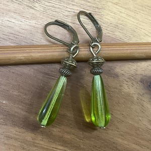 May include: A pair of drop earrings featuring translucent lime green glass beads in a teardrop shape. The beads are suspended from antique bronze-toned metal lever-back hooks, accented with small, decorative, textured metal spacer beads at the top.