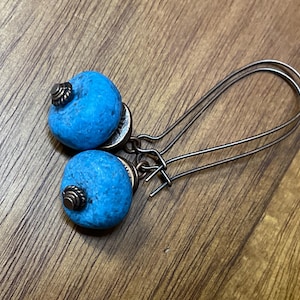 May include: A pair of drop earrings featuring round, textured, bright blue beads. Each bead is accented with a small, dark metallic cap and a coiled metal detail at the top. The earrings hang from long, curved, dark bronze-toned metal ear wires.