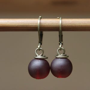 Puede incluir: Un par de pendientes colgantes con cuentas redondas de color rojo oscuro. Las cuentas cuelgan de ganchos para la oreja de color latón antiguo. Los pendientes se muestran en una clavija de madera sobre un fondo neutro.