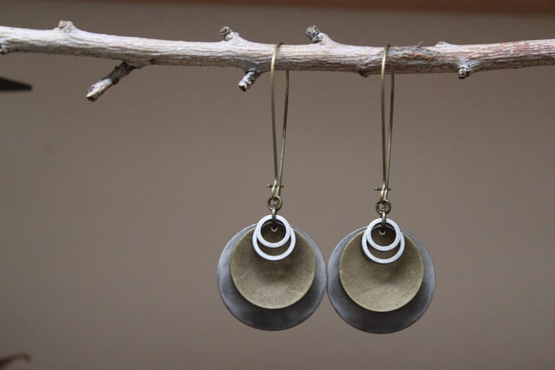 May include: A pair of dangle earrings featuring a layered design with a large dark grey circular disc, a smaller brass-toned circular disc, and two small silver-toned rings suspended from a long, thin, antique brass-colored metal ear wire hook.