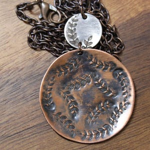 May include: A copper pendant necklace with a detailed leaf design. The necklace features a large, round pendant with an embossed leaf pattern and a smaller, silver pendant. The chain is a dark bronze colour.