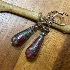 May include: A pair of drop earrings featuring teardrop-shaped beads with a mottled red and green pattern. The beads are accented with antique copper-toned metal spacers and attached to matching copper-toned leverback ear wires for secure closure.
