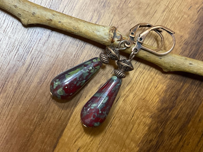 May include: A pair of drop earrings featuring teardrop-shaped beads with a mottled red and green pattern. The beads are accented with antique copper-toned metal spacers and attached to matching copper-toned leverback ear wires for secure closure.