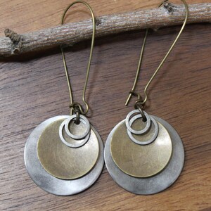 May include: A pair of dangle earrings featuring layered circular discs in brushed silver and brass tones. Each earring consists of a large silver disc, a smaller brass disc, and two tiny silver rings suspended from a curved brass ear wire hook.