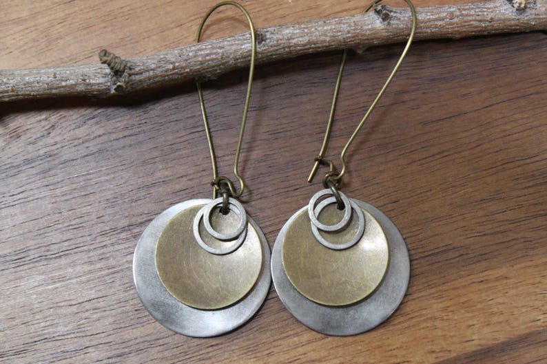 May include: A pair of dangle earrings featuring layered circular discs in brushed silver and brass tones. Each earring consists of a large silver disc, a smaller brass disc, and two tiny silver rings suspended from a curved brass ear wire hook.