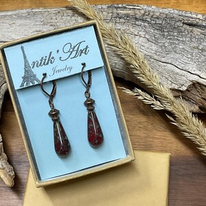 May include: A pair of drop earrings featuring dark red, speckled glass teardrop beads suspended from bronze-toned leverback hooks. The earrings are displayed in a rectangular box with a light blue card labeled Antik'Art Jewelry.
