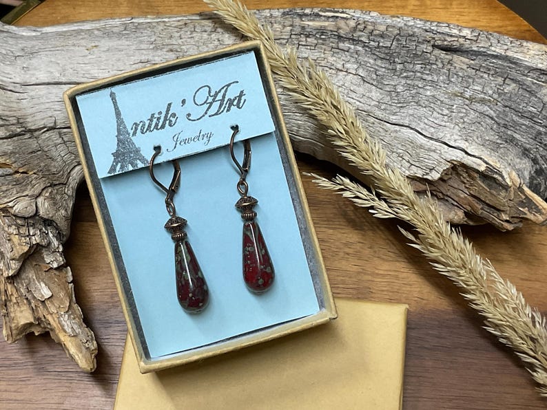 May include: A pair of drop earrings featuring dark red, speckled glass teardrop beads suspended from bronze-toned leverback hooks. The earrings are displayed in a rectangular box with a light blue card labeled Antik'Art Jewelry.