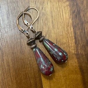 May include: A pair of drop earrings featuring teardrop-shaped beads with a mottled red and green glaze. The beads are suspended from antique copper-toned metal leverback hooks, accented by small, textured, conical metal spacers above each bead.
