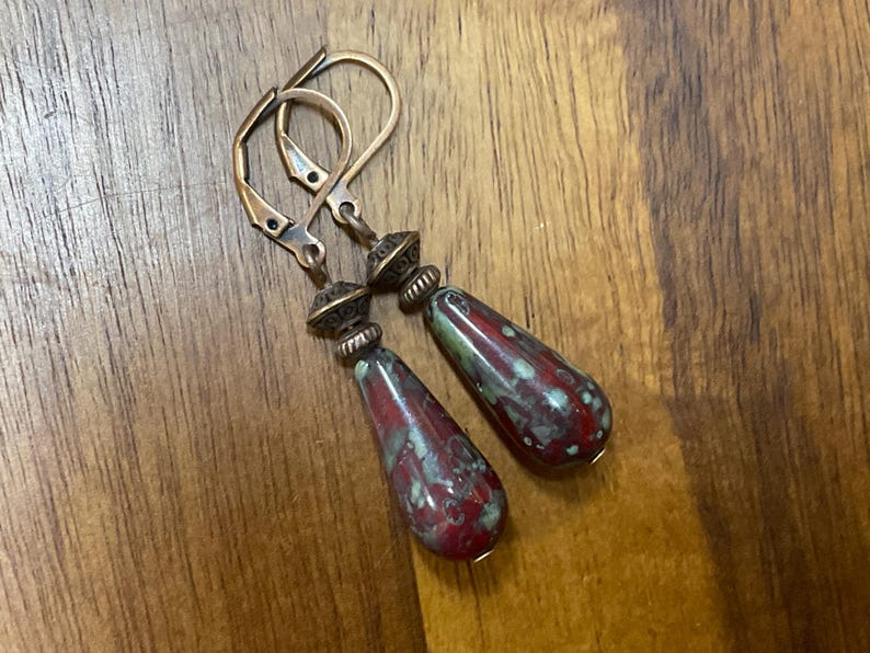 May include: A pair of drop earrings featuring teardrop-shaped beads with a mottled red and green glaze. The beads are suspended from antique copper-toned metal leverback hooks, accented by small, textured, conical metal spacers above each bead.