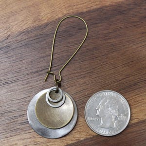 May include: A single earring featuring a long, curved bronze-toned wire hook attached to a layered pendant. The pendant consists of a large silver-toned disc, a smaller bronze-toned disc, and a tiny silver ring. A US quarter is shown for scale.