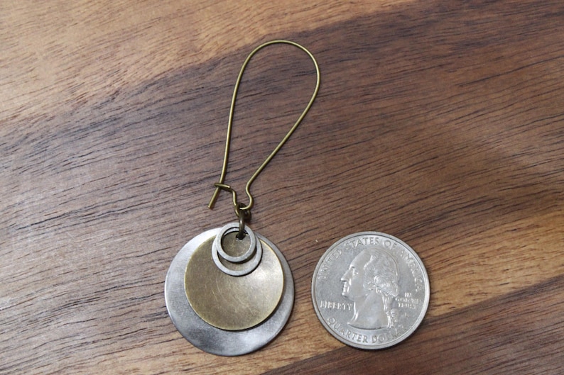 May include: A single earring featuring a long, curved bronze-toned wire hook attached to a layered pendant. The pendant consists of a large silver-toned disc, a smaller bronze-toned disc, and a tiny silver ring. A US quarter is shown for scale.