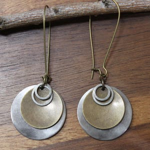 May include: A pair of dangle earrings featuring layered metal discs in brushed silver and antique brass tones. Each earring consists of a larger silver disc, a smaller brass disc, and a tiny silver ring, all suspended from a long brass hook wire.