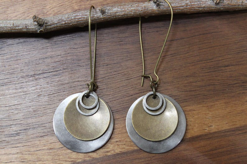 May include: A pair of dangle earrings featuring layered metal discs in brushed silver and antique brass tones. Each earring consists of a larger silver disc, a smaller brass disc, and a tiny silver ring, all suspended from a long brass hook wire.