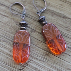 May include: A pair of drop earrings featuring elongated, translucent orange glass beads with an etched leaf pattern. Each bead is topped with a decorative, antiqued copper-toned metal bead and a secure lever-back ear wire in a matching finish.
