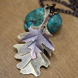 May include: A handcrafted leaf pendant necklace. The pendant features two overlapping metal leaves in shades of bronze, copper, and silver. The necklace is displayed on a turquoise stone and a wooden surface.