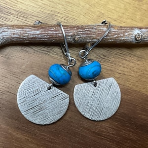 May include: A pair of drop earrings featuring silver-toned metal semi-circle charms with a brushed texture. Each earring is accented with a round, bright turquoise blue bead with dark veining, suspended from a silver-toned metal leverback hook.