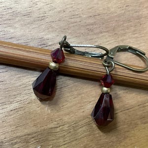 May include: A pair of drop earrings featuring deep red faceted crystal beads in a teardrop shape, accented with small gold-toned spacer beads. The earrings are attached to antique brass-colored lever-back closures, providing a secure fit.