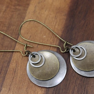 May include: A pair of dangle earrings featuring antique brass ear wires and a layered design. Each earring consists of a large silver-toned circular disc, a smaller brass-toned circular disc, and two small silver-toned rings suspended from the wire.