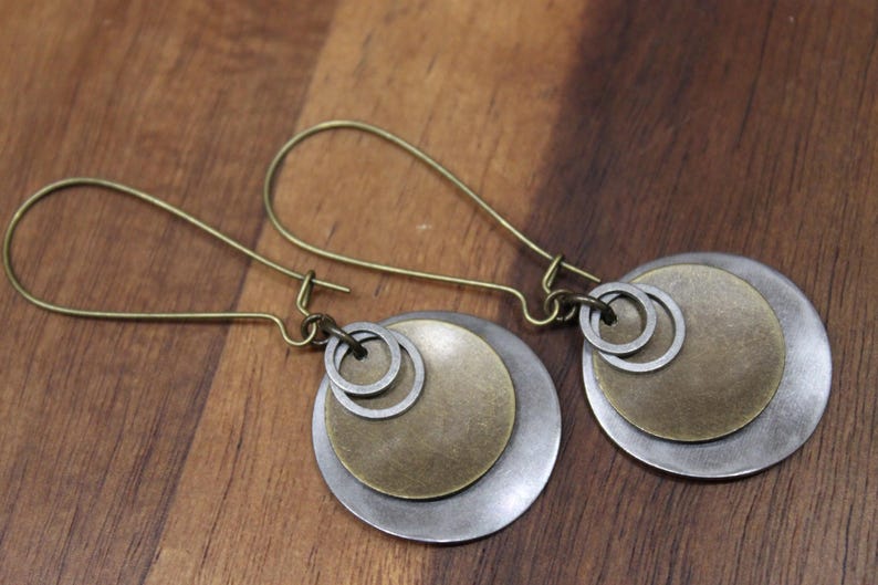May include: A pair of dangle earrings featuring antique brass ear wires and a layered design. Each earring consists of a large silver-toned circular disc, a smaller brass-toned circular disc, and two small silver-toned rings suspended from the wire.