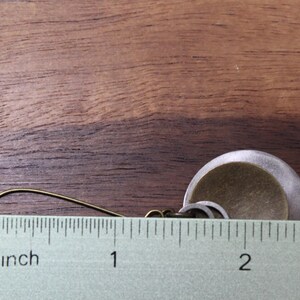 May include: A dangle earring featuring a brass-toned hook and a layered circular pendant composed of a smaller, textured brass-colored disc centered on a larger, matte silver-toned disc. A ruler shows the earring measures approximately 1.75 inches.