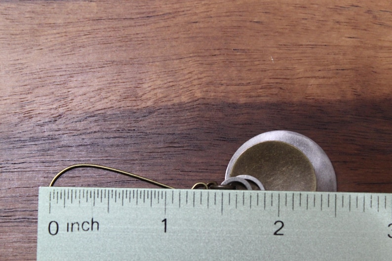 May include: A dangle earring featuring a brass-toned hook and a layered circular pendant composed of a smaller, textured brass-colored disc centered on a larger, matte silver-toned disc. A ruler shows the earring measures approximately 1.75 inches.