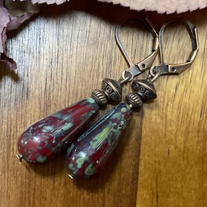 May include: A pair of drop earrings featuring elongated, teardrop-shaped beads with a mottled red and green glaze. Each bead is topped with a textured, antique-bronze-toned metal spacer and a matching lever-back ear wire for secure attachment.