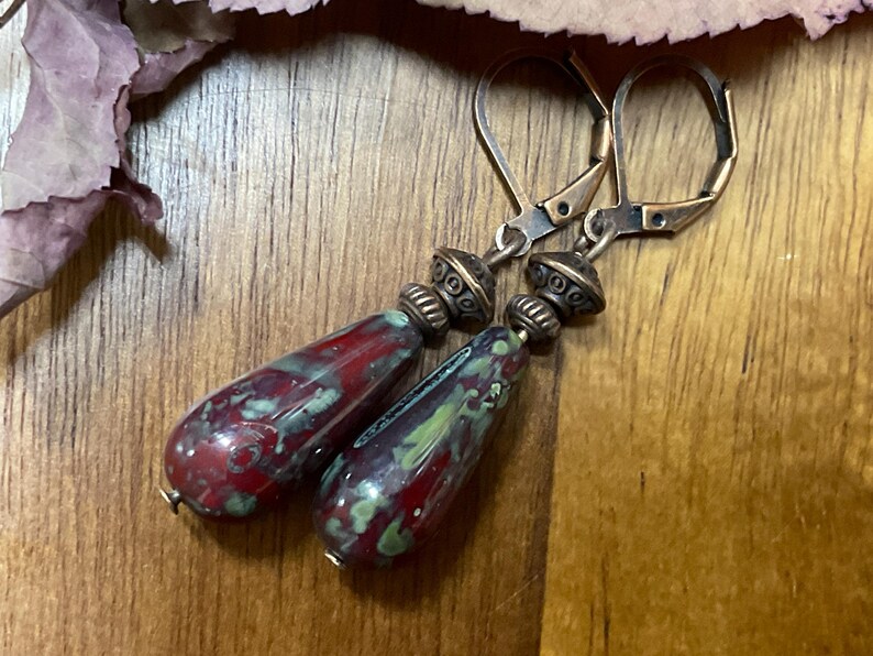 May include: A pair of drop earrings featuring elongated, teardrop-shaped beads with a mottled red and green glaze. Each bead is topped with a textured, antique-bronze-toned metal spacer and a matching lever-back ear wire for secure attachment.