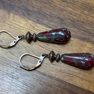 May include: A pair of drop earrings featuring teardrop-shaped beads with a mottled red and green glaze. Each bead is accented by two small, textured antique bronze-toned metal spacers and attached to a matching bronze-toned leverback ear wire.