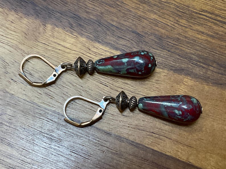 May include: A pair of drop earrings featuring teardrop-shaped beads with a mottled red and green glaze. Each bead is accented by two small, textured antique bronze-toned metal spacers and attached to a matching bronze-toned leverback ear wire.