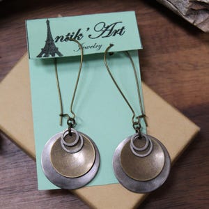 May include: A pair of dangle earrings featuring layered circular discs in antique brass and silver tones, suspended from long curved ear wires. The earrings are attached to a light green display card printed with the text Antik' Art Jewelry.