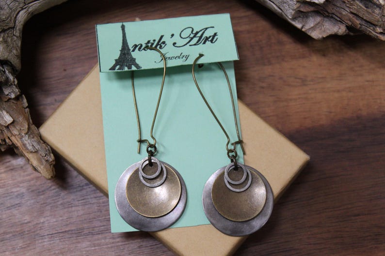 May include: A pair of dangle earrings featuring layered circular discs in antique brass and silver tones, suspended from long curved ear wires. The earrings are attached to a light green display card printed with the text Antik' Art Jewelry.