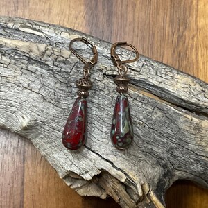 May include: A pair of drop earrings featuring teardrop-shaped beads with a mottled red and green glaze. The beads are suspended from copper-toned metal leverback hooks and accented with small, decorative, ridged copper-toned metal spacer beads.