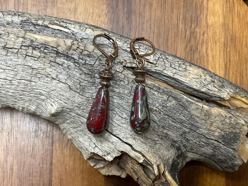 May include: A pair of drop earrings featuring teardrop-shaped beads with a mottled red and green glaze. The beads are suspended from copper-toned metal leverback hooks and accented with small, decorative, ridged copper-toned metal spacer beads.