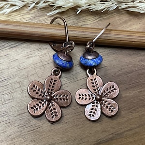 Op de afbeelding: Een paar oorbellen van koperkleurig metaal met een vijfbladige bloemhanger met geëtste bladachtige details. Elke oorbel is voorzien van een kleine, ronde, felblauwe stenen kraal die boven de bloemhanger op een klapoorbelhaak is geplaatst.