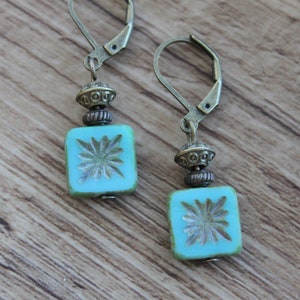 May include: A pair of turquoise square earrings with a floral design, accented with antique brass-colored metal beads and lever-back closures. The earrings are set against a wooden background.