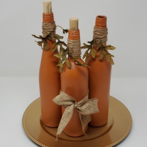 Puede incluir: Tres botellas de vidrio naranja con tapas de corcho y detalles de cuerda de yute, decoradas con hojas de oto&ntilde;o y un lazo de yute, sobre un plato dorado.