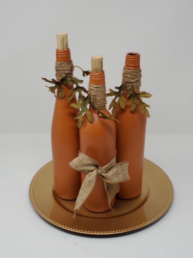 Puede incluir: Tres botellas de vidrio naranja con tapas de corcho y detalles de cuerda de yute, decoradas con hojas de oto&ntilde;o y un lazo de yute, sobre un plato dorado.